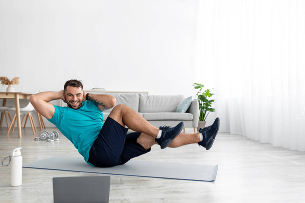 Joven atleta europeo alegre en uniforme haciendo ejercicios abdominales en la estera con el ordenador portátil en la sala de estar - Foto, imagen