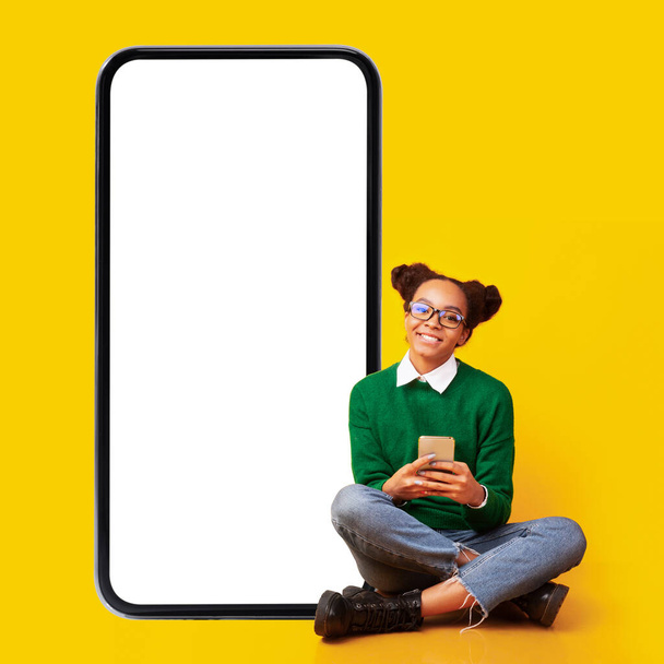 Adolescente africaine utilisant le téléphone assis près du smartphone, fond jaune - Photo, image