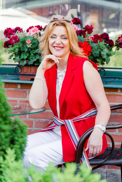 Een mooie volwassen vrouw in een wit en rood pak zit op een zomerterras. Vrouw gelukkig leven concept na 40 jaar - Foto, afbeelding