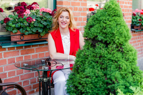 Krásná zralá žena v bílém a červeném obleku sedí na letní terase. Žena šťastný život koncept po 40 letech - Fotografie, Obrázek