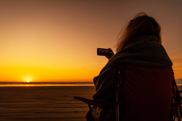 Femme caucasienne couverte d'une couverture chaude prenant la vidéo d'un coucher de soleil sur une plage panoramique alors qu'elle était assise sur une chaise de camp. Front de mer vacances thème. - Photo, image