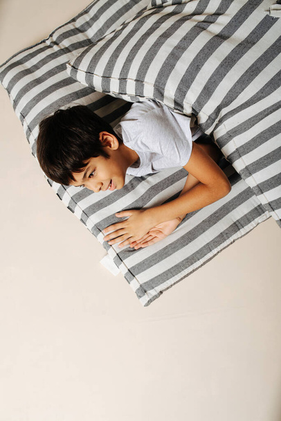Animated indian boy looking at someone from under a blanket. Lying on a mattress on the floor, over beige. - Fotografie, Obrázek