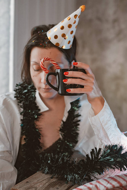Väsynyt nainen puolueen hattu krapula ja päänsärky juhlimisen jälkeen uudenvuodenaattona istuu silmät kiinni ja pää alas puhdistamaton juhlapöytä kotona, kärsivät migreeni ja unen puute - Valokuva, kuva