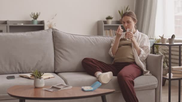 Puukota hidas laukaus hymyilevä nuori valkoihoinen raskaana oleva nainen ottaa synnytystä vitamiineja istuu sohvalla kotona - Materiaali, video