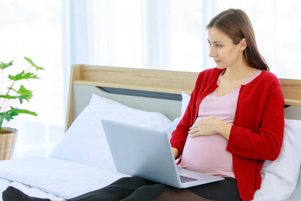 biały młody szczęśliwy zdrowe kobieta prenatalny ciężarna matka model w casual strój z kurtka siedzi na łóżko w sypialnia trzymając rękę na duży brzuch oglądać śmieszne komedia film za pośrednictwem laptop komputer. - Zdjęcie, obraz