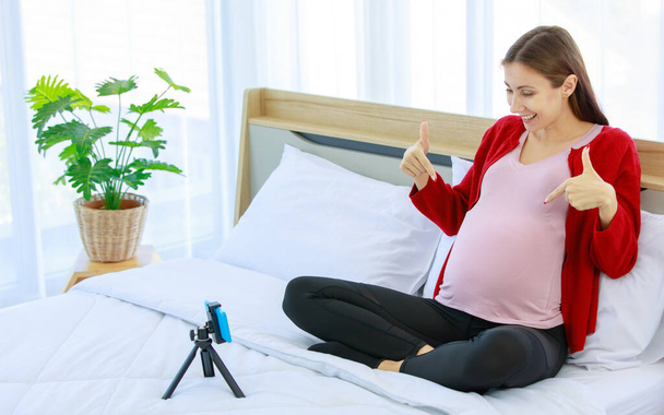 Blanc millénaire jeune heureux sain femme enceinte prénatale modèle mère en tenue décontractée assis souriant sur le lit dans la chambre à coucher tenant la main sur le gros ventre avec amour en utilisant smartphone appel vidéo. - Photo, image