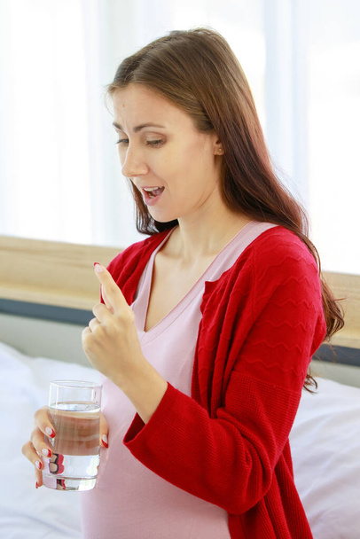 Closeup shot of Caucasian millennial young female prenatal pregnant mother in casual pregnancy outfit with big belly tummy sitting holding vitamin healthcare medicine pill and drinking water glass. - Valokuva, kuva