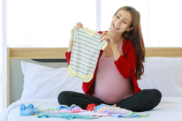 Caucasian millennial young happy female prenatal pregnant mother in casual pregnancy outfit jacket sitting smiling on bed in bedroom holding preparing choosing small cotton baby clothes in hands. - Foto, Bild