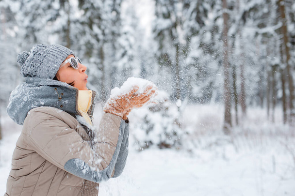 šťastná žena kráčí venku v zasněženém zimním dni. Žena model oblečený módní šedá nosit sluneční brýle foukání sníh baví, chytání sněhu s rukama - Fotografie, Obrázek