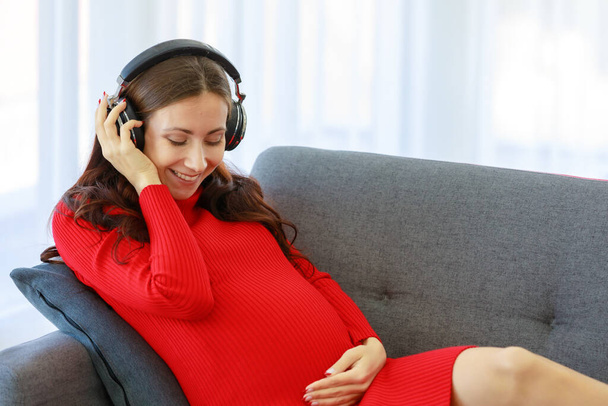 白人千年紀若いです幸せなセクシーな女性妊娠中の母でカジュアル赤妊娠ドレスダウン上の居心地の良いソファ身に着けていますヘッドフォン音楽プレイリストに耳を傾けます笑顔保持手上の腹. - 写真・画像