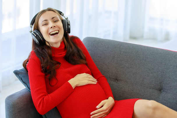 白人千年紀若いです幸せなセクシーな女性妊娠中の母でカジュアル赤妊娠ドレスダウン上の居心地の良いソファ身に着けていますヘッドフォン音楽プレイリストに耳を傾けます笑顔保持手上の腹. - 写真・画像