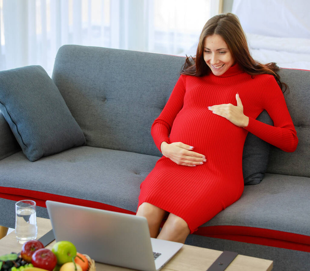 kaukaskie millennial młody szczęśliwy sexy kobieta prenatal ciąży matka w casual czerwony ciąży sukienka siedzi na kanapie trzymając się za ręce na brzuchu z miłości za pomocą laptopa przeglądania Internetu. - Zdjęcie, obraz