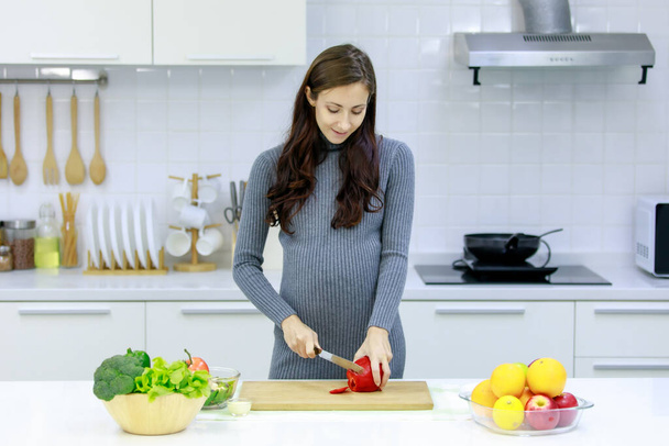 Кавказский тысячелетие молодая счастливая сексуальная женщина беременная мать в случайном сером платье беременности стоя улыбаясь с помощью ножа резки красный сладкий перец на резке доски приготовления овощей салат. - Фото, изображение