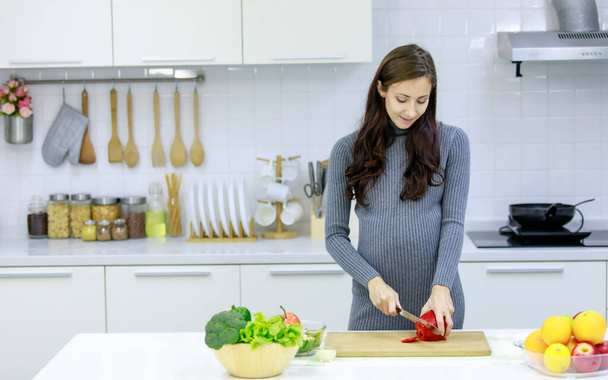 Kaukázusi Y-generációs fiatal boldog szexi nő prenatal terhes anya alkalmi szürke terhesség ruha áll mosolyogva késsel vágás piros édes bors vágódeszka főzés zöldség saláta. - Fotó, kép