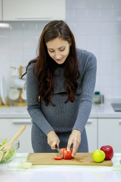 Kaukázusi Y-generációs fiatal boldog szexi nő prenatal terhes anya alkalmi szürke terhesség ruha áll mosolyogva késsel vágás piros édes bors vágódeszka főzés zöldség saláta. - Fotó, kép