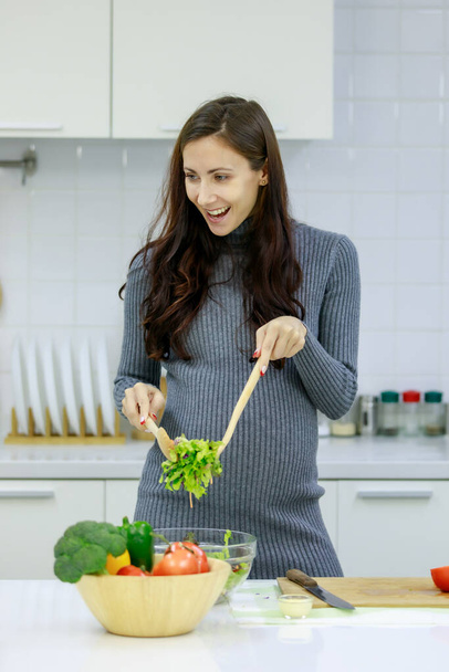 Blanc millénaire jeune heureux sexy femme enceinte prénatale mère en robe de grossesse grise occasionnelle debout souriant à l'aide d'un couteau coupe poivron rouge sur planche à découper cuisine légumes salade. - Photo, image