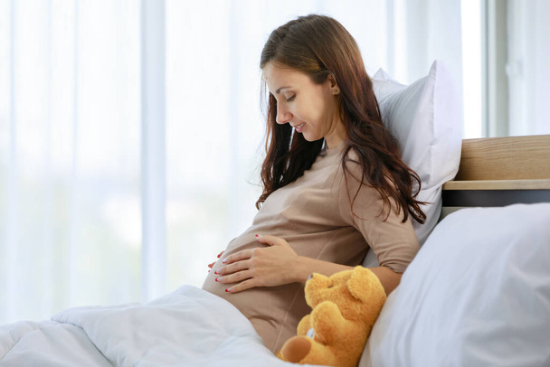 Caucasian millennial young happy female prenatal pregnant mother in casual shirt sitting smiling under blanket on bed in bedroom holding hands touching baby in big belly tummy with love and care. - Фото, изображение