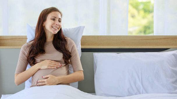 Caucasien millénariste jeune mère enceinte prénatale heureuse en chemise décontractée assis souriant sous la couverture sur le lit dans la chambre à coucher tenant les mains touchant bébé dans le gros ventre avec amour et soin. - Photo, image