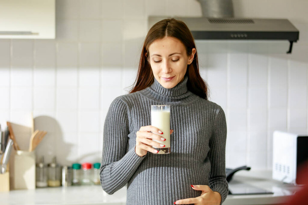 Caucasian millennial young happy female prenatal pregnant mother in casual outfit standing alone holding hand on big belly tummy with love drinking fresh milk for breakfast in full equipment kitchen. - Photo, Image
