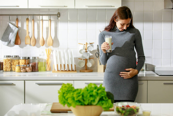 Caucasian millennial young happy female prenatal pregnant mother in casual outfit standing alone holding hand on big belly tummy with love drinking fresh milk for breakfast in full equipment kitchen. - Foto, Bild