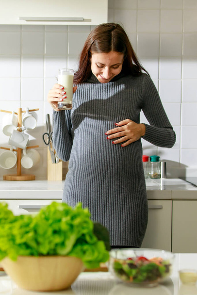 Caucasian millennial young happy female prenatal pregnant mother in casual outfit standing alone holding hand on big belly tummy with love drinking fresh milk for breakfast in full equipment kitchen. - Photo, Image