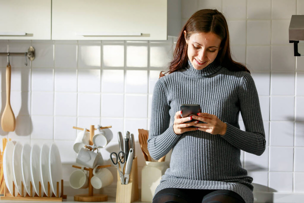 Caucasian millennial young happy female prenatal pregnant mother in casual dress standing smiling holding smartphone in hand taking selfie photo, greeting say hi video call with family in kitchen. - Fotoğraf, Görsel