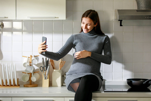 kaukaskie millennial młody szczęśliwy kobieta prenatal ciąży matka w casual sukienka stoi uśmiechnięty gospodarstwa smartfon w ręku biorąc selfie zdjęcie, pozdrowienia powiedzieć cześć wideo połączenie z rodziną w kuchni. - Zdjęcie, obraz