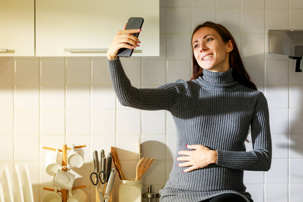 Καυκάσιος χιλιετή νεαρή ευτυχισμένη γυναίκα προγεννητική έγκυος μητέρα σε περιστασιακό φόρεμα στέκεται χαμογελώντας κρατώντας smartphone στο χέρι λήψη selfie φωτογραφία, χαιρετισμό πω γεια video call με την οικογένεια στην κουζίνα. - Φωτογραφία, εικόνα