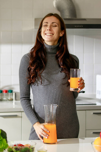 Caucasian millennial young happy female prenatal pregnant mother in casual outfit standing alone holding hand on big belly tummy with love drinking orange juice for breakfast in full equipment kitchen. - Foto, afbeelding