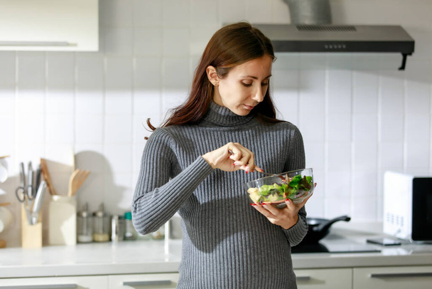 Caucasian millennial young happy sexy female prenatal pregnant mother in casual gray pregnancy dress sitting smiling using fork eating red tomato in vegetables salad bowl in full equipment kitchen. - 写真・画像