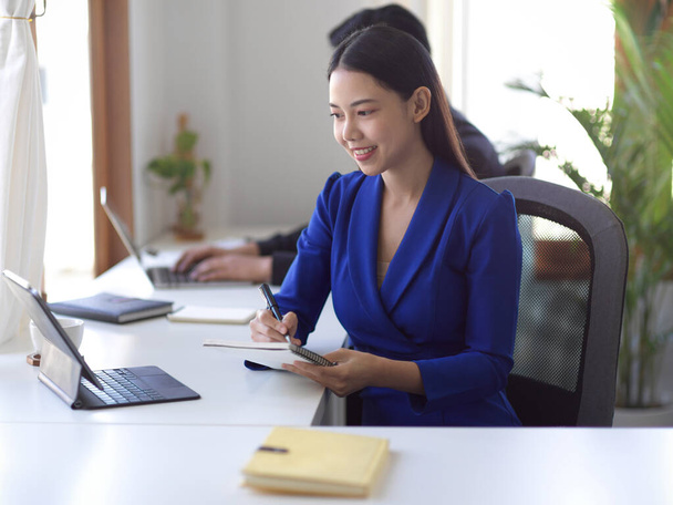幸せなアジアの女性実業家は、タブレット画面を見て、オフィスの職場で机の上にメモを取ります. - 写真・画像