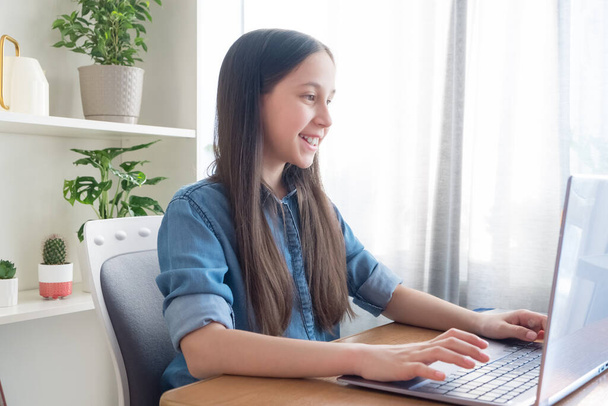Een brunette gekleed in een denim shirt studeert thuis aan de computer, studeert, doet haar huiswerk, communiceert op het netwerk - Foto, afbeelding
