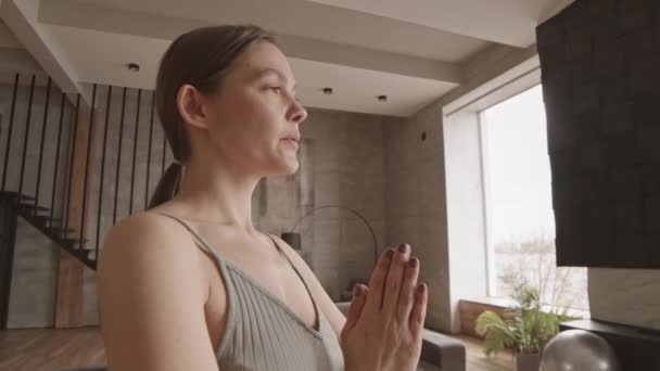 Handheld slowmo πλάνο της νεαρής Καυκάσιας γυναίκας στέκεται με τα χέρια προσεύχεται και κοιτάζοντας ευθεία μπροστά, ενώ η πρακτική γιόγκα στο σπίτι που ζουν στο σύγχρονο διαμέρισμα διπλής όψης - Πλάνα, βίντεο