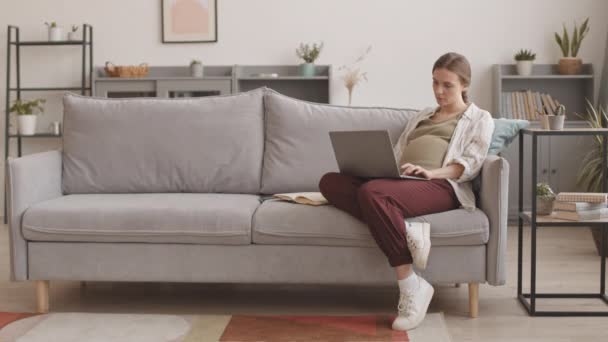Медленный удар ножом снимок молодой белой беременной женщины, работающей на ноутбуке из дома, сидящей в расслабленном положении на диване в гостиной - Кадры, видео