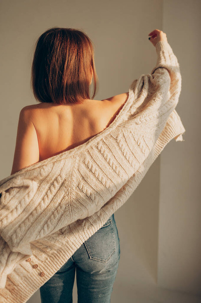 sexy young woman with bare shoulders in knitted cardigan, jeans moves freely in sunlight in white bright room. women's dances and movements. vertically, selective focus - Fotografie, Obrázek