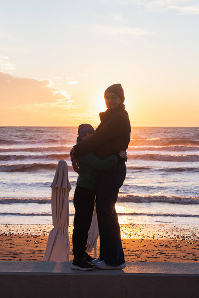 Mom and son are enjoying the sunset over the sea with a beautiful horizon. Wonders of nature. Beautiful landscape of the winter sea. - Photo, Image