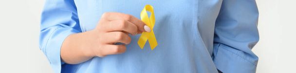 Female nurse holding golden awareness ribbon on light background, closeup. International Childhood Cancer Day - Photo, Image