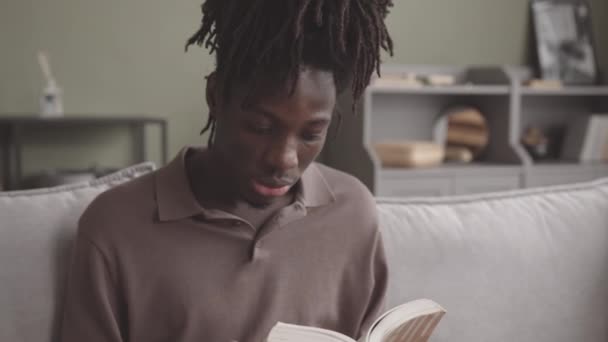 Zoom-out slowmo shot van geconcentreerde jonge zwarte man voorlezen hardop zitten op de bank thuis - Video