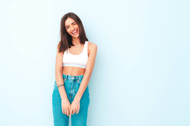 Portrait of beautiful smiling woman dressed in jeans. Sexy carefree cheerful model. Adorable and positive female posing near light blue wall in studio. Tanned and skinny. Shows tongue - Photo, image