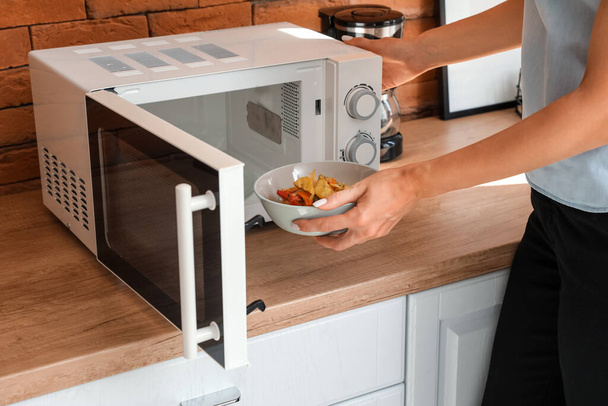 Woman putting bowl with delicious food into modern microwave oven on wooden counter near brick wall - Photo, Image