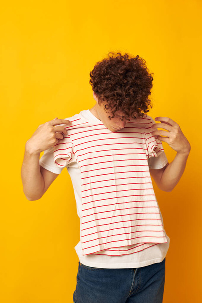 guy with red curly hair in a white t-shirt holds a striped t-shirt fashion youth style yellow background unaltered - Photo, image