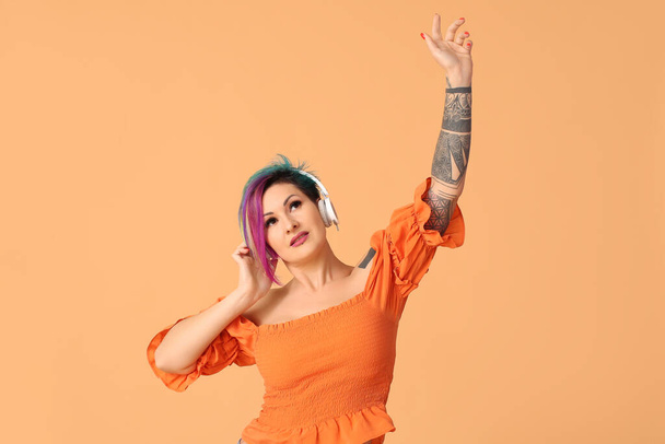 Beautiful woman with unusual hair and tattoo listening to music on orange background - Photo, image