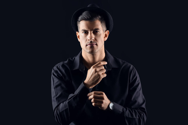 Fashionable gentleman in stylish hat adjusting sleeve on dark background - Valokuva, kuva