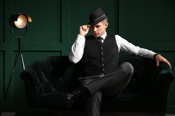 Джентльмен в формальной одежде и стильной шляпе сидит на диване напротив цветной стены - Фото, изображение