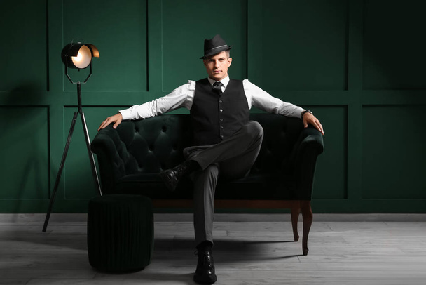 Κύριος με επίσημα ρούχα και κομψό καπέλο κάθεται σε καναπέ στο σκοτεινό δωμάτιο - Φωτογραφία, εικόνα