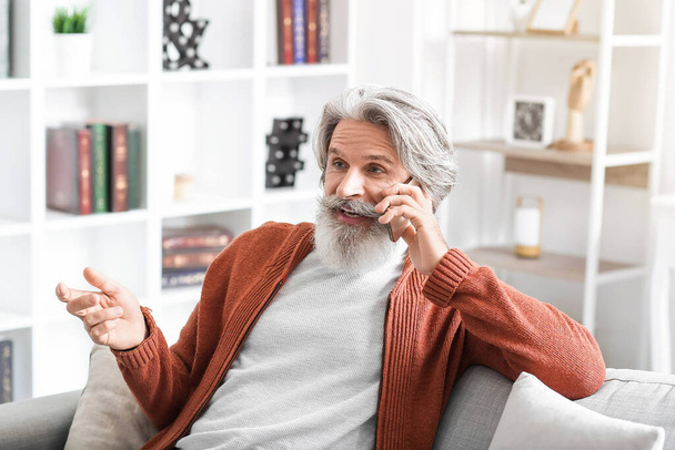 Senior man praat met mobiele telefoon op de bank thuis - Foto, afbeelding