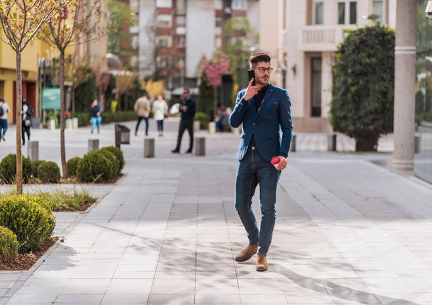 Homme d'affaires urbain en manteau bleu marchant dans la ville et parlant sur téléphone portable - Photo, image