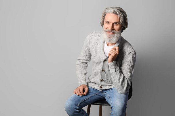 Hezký starší muž v pleteném svetru sedí na židli proti šedému pozadí - Fotografie, Obrázek