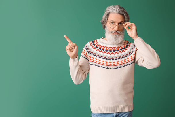 Jóképű idős férfi kötött pulóverben, aki valami zöld hátterűre mutat. - Fotó, kép