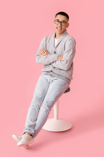 Красивый молодой азиат в вязаном свитере сидит на стуле на розовом фоне - Фото, изображение